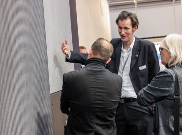 Ein Fachberater erläutert zwei Besuchern auf einer Messe die Details einer strukturierten Faserzementtafel – im Fokus stehen Materialität, Haptik und Anwendungsmöglichkeiten für vorgehängte hinterlüftete Fassaden.