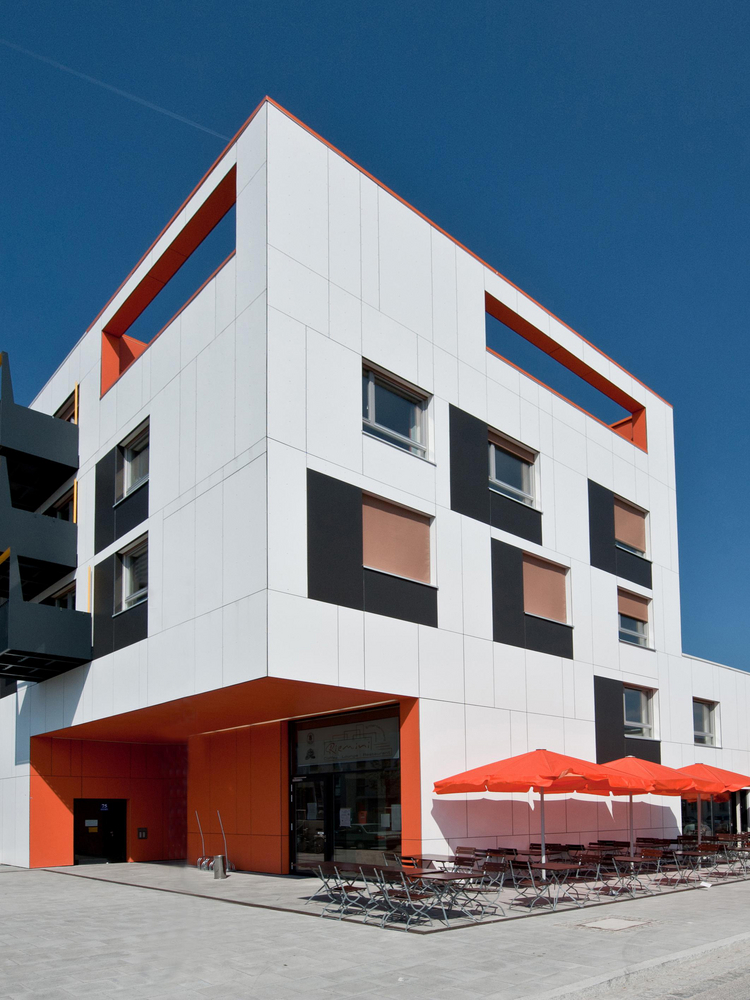 Markantes Gebäude mit Fassade aus großformatigen EQUITONE Faserzementtafeln in Weiß, akzentuiert durch schwarze und orangefarbene Flächen – die kontrastreiche Farbgebung und geometrische Gestaltung verleihen dem Bau eine starke Identität und betonen die Vielseitigkeit vorgehängter hinterlüfteter Fassadensysteme.