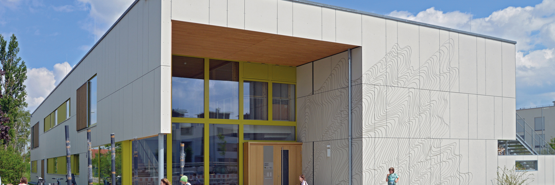 Zweigeschossiges, modern gestaltetes Gebäude mit heller Fassade aus Faserzementtafeln. Die Tafeln sind mit einer filigranen Gravurstruktur versehen. Große Fensterflächen und ein überdachter Eingangsbereich mit gelbgrünen Rahmen betonen die Offenheit des Gebäudes. Kinder spielen vor dem Gebäude, was auf eine Nutzung als Bildungseinrichtung (z. B. Kindergarten oder Schule) hinweist. Der Himmel ist klar und blau, was die freundliche Atmosphäre unterstreicht.