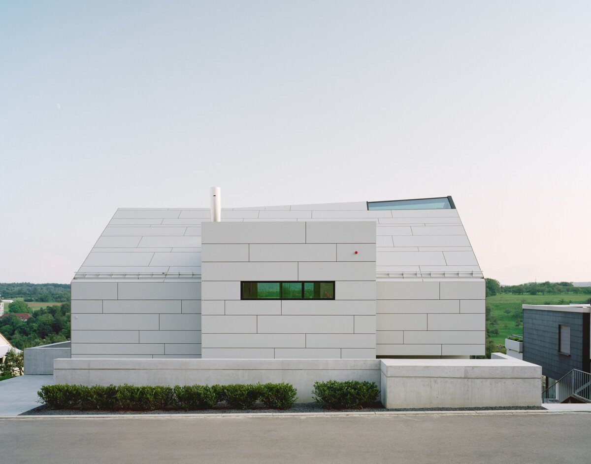 Wohnhaus mit durchgängigem Systemdach aus hellen Faserzementtafeln, das Dach- und Fassadenflächen nahtlos miteinander verbindet; die horizontale Linienstruktur und präzise Plattenanordnung unterstreichen die moderne Architektursprache und klare Geometrie des Baukörpers.